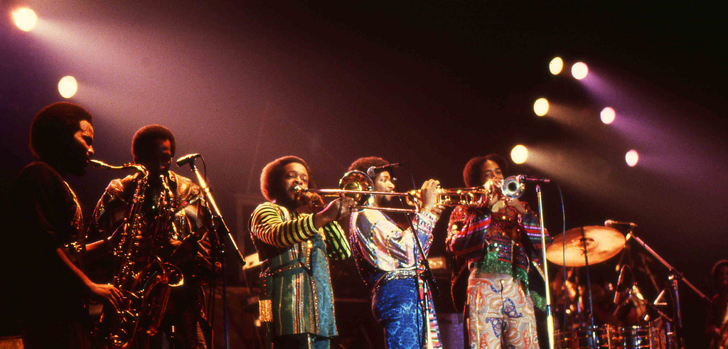 Earth, Wind & Fire: Uma Odisseia Biográfica de Alma, Funk e Espiritualidade
1. Introdução: O Som Inconfundível dos Elementos
Earth, Wind & Fire (EWF) irrompeu na paisagem musical com uma fusão sonora tão inovadora que estava destinada a ser imitada por muitos, mas nunca duplicada com sucesso por ninguém. Desde o início, a banda transcendeu as fronteiras de gênero e raça, tornando-se um dos primeiros grupos negros a alcançar sucesso massivo com o público branco e, ao fazê-lo, abriu caminho para o fenômeno do crossover que definiria as décadas seguintes. Sua ascensão não foi apenas um sucesso comercial; foi uma redefinição cultural.
Nos anais da música dos anos 1970, o EWF ocupa um lugar singular. Seu som era uma alquimia magistral de R&B, rock, soul, funk e ritmos vibrantes da África e da América Latina, construído sobre uma base de funk dançante, arranjos de metais exuberantes e o falsete etéreo de Philip Bailey. No entanto, o que solidificou seu apelo duradouro foi sua filosofia: uma mensagem de positividade, amor e misticismo universal que convidava os ouvintes a se conectarem com algo maior. Essa combinação de sofisticação musical e profundidade espiritual criou uma força inegável.
Por trás dessa visão sonora complexa, mas unificada, estava a genialidade de uma figura central: o fundador, líder e guia espiritual do grupo, Maurice White.
2. As Origens: A Visão Formativa de Maurice White
Maurice White foi a força motriz e o coração filosófico do Earth, Wind & Fire. Mais do que um músico, ele foi um arquiteto sonoro cuja visão foi moldada por uma jornada musical rica e diversificada. Suas primeiras experiências, desde os corais gospel de Memphis até os prestigiosos estúdios de gravação de Chicago, foram cruciais para sintetizar o som e a identidade únicos da banda que ele viria a criar.
A jornada musical de White foi uma preparação metódica para sua obra-prima:
* Formação Inicial: Nascido em Chicago e criado em Memphis, a educação musical de White começou aos seis anos em um coral gospel. Seu fascínio juvenil por bandas marciais o levou à bateria, o instrumento que se tornaria sua primeira porta de entrada para o mundo profissional.
* Desenvolvimento Profissional: Após o ensino médio, White retornou a Chicago para estudar no Conservatório de Música. Simultaneamente, ele se estabeleceu como um requisitado baterista de sessão na lendária Chess Records, onde, ao longo dos anos 1960, colaborou com ícones como Muddy Waters e The Impressions, absorvendo a essência do blues e da soul music.
* A Influência de Ramsey Lewis: De 1967 a 1969, o período de White no aclamado Ramsey Lewis Trio provou ser uma influência primordial em sua filosofia musical. Ele revelou à Billboard seu desejo de criar algo que seguisse os passos de Lewis: "algo que fosse musical, mas ainda assim divertido, e algo em que o tema tocasse seu coração". Foi Lewis quem o apresentou à kalimba (o piano de polegar africano), cujo som místico se tornaria uma assinatura sonora indelével do EWF.
Com essa visão artística amadurecida, White sentiu a necessidade de criar seu próprio veículo musical, uma ambição que o levaria a Los Angeles para materializar seu sonho.
3. A Formação da Banda: Dos Salty Peppers ao Renascimento Astrológico
Deixando para trás a segurança de ser um músico acompanhante de elite, Maurice White mudou-se para Los Angeles em 1969 com um objetivo estratégico claro: montar um grande conjunto musical autossuficiente. Sua visão era criar uma banda com um elenco fixo de talentos excepcionais, eliminando a dependência da contratação de músicos locais durante as turnês.
A evolução do grupo foi rápida e decisiva:
1. The Salty Peppers: A primeira encarnação da banda, chamada The Salty Peppers, assinou com a Capitol Records, mas, após alguns anos, não conseguiu alcançar notoriedade significativa.
2. O Nascimento do Earth, Wind & Fire: Em 1971, White garantiu um novo contrato com a Warner Brothers e rebatizou o grupo. O nome "Earth, Wind & Fire" foi extraído diretamente dos elementos primários de seu mapa astrológico, um reflexo da dimensão espiritual que ele pretendia infundir em sua música.
3. Primeiros Álbuns e Reformulação Estratégica: Sob o selo da Warner Bros., a banda lançou três álbuns em menos de dois anos — Earth, Wind & Fire, The Need of Love e a trilha sonora Sweet Sweetback’s Baadasss Song — e emplacou os singles "Love is Life" (1971) e "Evil" (1973). Insatisfeito, White tomou a decisão crucial em 1972 de mudar para a Columbia Records e reformular completamente a banda. Ele montou uma formação mais jovem e dinâmica que incluía o vocalista Philip Bailey, a cantora Jessica Cleaves (do The Friends of Distinction), o guitarrista Johnny Graham e o guitarrista Al McKay.
Essa nova e revitalizada formação, agora apoiada pela liberdade criativa da Columbia, estava prestes a desencadear a revolução sônica e visual que definiria a década seguinte.
4. A Era de Ouro: Sucesso, Inovação e Superestrelato (1972-1983)
A ascensão do Earth, Wind & Fire na década de 1970 não foi um mero acaso, mas a confluência magistral da visão de White, uma formação musicalmente virtuosa e o zeitgeist cultural da época. A era disco, em particular, serviu como um catalisador que os transformou em verdadeiros superstars. No entanto, enquanto muitos artistas foram definidos e limitados pelo gênero, o EWF transcendeu-o. Sua sofisticação harmônica e rítmica, enraizada no jazz e no soul, permitiu que eles dominassem a era sem serem confinados por ela.
Os marcos deste período dourado ilustram seu domínio e inovação:
* O Ponto de Virada: O avanço definitivo para o sucesso mainstream veio em 1975 com a trilha sonora do filme That's the Way of the World. O álbum alcançou o topo das paradas, impulsionado pelo single "Shining Star", que não apenas liderou as paradas de pop e R&B, mas também rendeu à banda seu primeiro prêmio Grammy.
* Domínio nas Paradas: O sucesso tornou-se a norma. Entre 1971 e 1988, o EWF emplacou 38 hits nas paradas de R&B, com a grande maioria (32) cruzando para as paradas pop. Álbuns de ouro, platina e platina dupla se seguiram, solidificando seu status como uma potência comercial.
* Inovação nos Palcos: Nos palcos, o EWF transcendia o conceito de um concerto; suas turnês em estádios eram rituais sônicos e visuais. Eles usavam os "locais cavernosos" a seu favor, criando espetáculos teatrais com ilusões do mestre Doug Henning. Pirâmides gigantes, fogos de artifício, truques de desaparecimento e a visão da banda "voando" para o palco criavam uma experiência mística inesquecível.
* Sucessos Icônicos: Esta era produziu um fluxo contínuo de hinos atemporais, incluindo:
* "Got to Get You into My Life" (um cover dos Beatles que se tornou um sucesso no Top 10)
* "September"
* "Sing a Song"
* "Boogie Wonderland" (com a participação do grupo The Emotions)
Após uma década de sucesso implacável e turnês exaustivas, a banda sentiu a necessidade de uma pausa, sinalizando uma mudança de ritmo em sua jornada monumental.
5. Hiato e Empreendimentos Individuais (1983-1986)
Em 1983, o Earth, Wind & Fire anunciou oficialmente um hiato. Longe de ser um fim, esta foi uma fase de diversificação artística que permitiu aos seus membros principais expandir seus horizontes e solidificar legados individuais. Maurice White e Philip Bailey, em particular, floresceram, demonstrando a profundidade do talento que impulsionava a banda.
Maurice White: O Produtor Visionário
Maurice White aproveitou o hiato para fundar sua empresa, a Kalimba Productions, e se firmar como um produtor e compositor de elite. Seu toque de Midas foi procurado por uma lista impressionante de artistas, para os quais ele produziu álbuns aclamados, incluindo Barbra Streisand, Neil Diamond, seu mentor Ramsey Lewis e The Emotions. Seu trabalho também se estendeu ao cinema, com contribuições para trilhas sonoras de filmes como Um Príncipe em Nova York (Coming to America).
Philip Bailey: O Sucesso Solo e a Fé
Philip Bailey lançou uma carreira solo de grande sucesso. Seu maior triunfo comercial foi o hit global "Easy Lover", um dueto icônico com Phil Collins que dominou as paradas mundiais. Paralelamente, Bailey explorou suas raízes na música gospel, recebendo aclamação da crítica e um prêmio Grammy pelo álbum Triumph!, consolidando sua reputação como um vocalista de extraordinária versatilidade e convicção.
Após essa pausa enriquecedora, os membros se prepararam para se reunir, enfrentando o desafio de reacender sua magia coletiva em um cenário musical que já havia se transformado.
6. Reuniões e Carreira Posterior: Navegando por Novas Décadas
O retorno do Earth, Wind & Fire, começando com o álbum Touch the World de 1987, foi marcado pela ambição de honrar seu legado enquanto navegava por uma paisagem musical em constante mudança. Novos gêneros haviam surgido, e a banda buscou encontrar seu lugar sem comprometer a identidade sônica e espiritual que os definia.
A trajetória da banda a partir do final dos anos 80 reflete tanto a resiliência quanto os desafios:
1. Os Primeiros Retornos: Os álbuns de reunião Touch the World (1987) e Heritage (1990) geraram hits nas paradas de R&B, mas tiveram um impacto pop mais modesto, um sinal das dificuldades em replicar o massivo apelo crossover de sua era de ouro.
2. O Legado em Retrospectiva: Em 1992, o lançamento do box set The Eternal Dance serviu como um documento histórico. Esta retrospectiva inovadora de 55 faixas cobriu duas décadas de música, incluindo raridades e faixas inéditas. O crítico James Hunter, na Musician, declarou que a coleção continha "algumas das músicas pop mais fluentes, centradas e elásticas já gravadas" e que "sua influência está em toda parte".
3. A Fase Millennium e Desafios com Gravadoras: O álbum Millennium (1993) marcou um retorno à Warner Bros., parcialmente motivado pelo descontentamento de Maurice White com a pressão da Columbia Records para incluir artistas convidados, como M.C. Hammer e Sly Stone, o que ele sentia que comprometia sua visão. Embora Millennium tenha sido aclamado por seu som clássico do EWF, as vendas foram decepcionantes.
Apesar dos desafios comerciais em suas décadas posteriores, o legado da banda já estava cimentado na história, transcendendo as flutuações do mercado e preparando o terreno para uma avaliação de seu impacto indelével.
7. Legado e Influência Duradoura: A Dança Eterna
O legado do Earth, Wind & Fire transcende as paradas de sucesso; eles se firmaram como uma força cultural e musical pioneira cujo impacto ressoa através de gerações. Sua música era "liricamente positiva, muitas vezes ousadamente espiritual", promovendo uma mensagem de "irmandade espiritual" que quebrava barreiras. Com sua imagem Afrocentric e som universal, eles criaram uma trilha sonora de esperança que permanece vital. Como disse o executivo musical Harry Weigner, "Ao longo de três décadas, Earth, Wind & Fire exibiram grooves deliciosos, musicalidade ousada, arranjos de tirar o fôlego, virtuosismo energético e um canto belíssimo."
O legado multifacetado da banda é evidente em suas contribuições duradouras:
* Impacto Crossover e Cultural: Como uma das primeiras bandas negras a conquistar um público branco em grande escala, o EWF alterou permanentemente a paisagem da música pop, abrindo caminho para que inúmeros artistas negros alcançassem o sucesso mainstream.
* Influência Musical Abrangente: Sua fusão de gêneros deixou uma marca indelével. Artistas como Prince, Michael Jackson, Beyoncé e Bruno Mars os citaram como uma influência fundamental. Seus grooves foram amplamente amostrados por artistas de hip-hop, garantindo sua relevância para novas gerações.
* Reconhecimento da Indústria: A excelência do grupo foi celebrada com os maiores prêmios da indústria, incluindo múltiplos Grammys, o NAACP Hall of Fame Image Award (1994) e a prestigiosa indução ao Rock & Roll Hall of Fame em 2000.
* Sucesso Comercial Monumental: Com mais de 90 milhões de álbuns vendidos em todo o mundo, o EWF alcançou um nível de sucesso global que comprova o poder duradouro e o apelo universal de sua música.
Earth, Wind & Fire: Biografia Musical